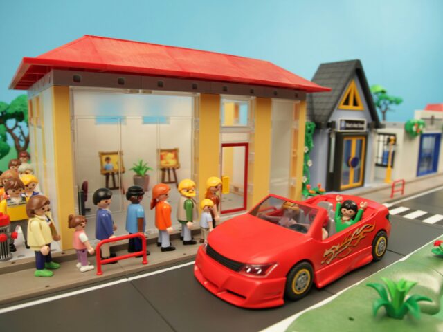 赤色の屋根の建物の横に駐車する赤い車とそれを見ている並んでいる人々