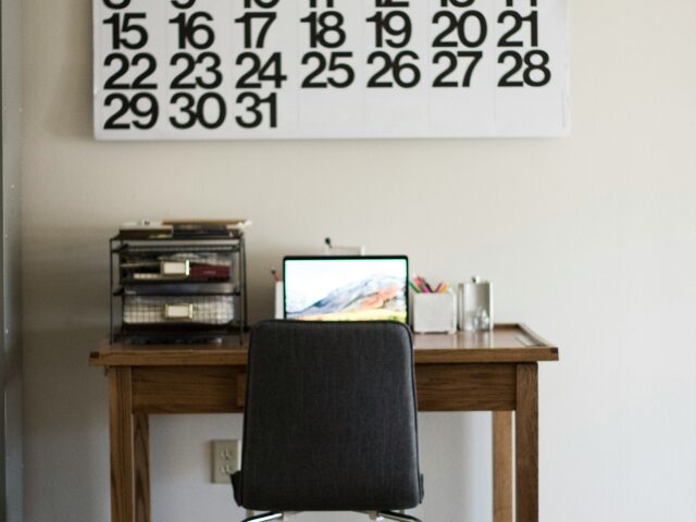 カレンダーが壁に貼ってありノートパソコンや書類が置いてあるお仕事机とイス